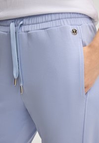 Close-up van een hand in een lichtblauwe elastische tailleband broekzak met trekkoord en een klein rond metalen logo detail.