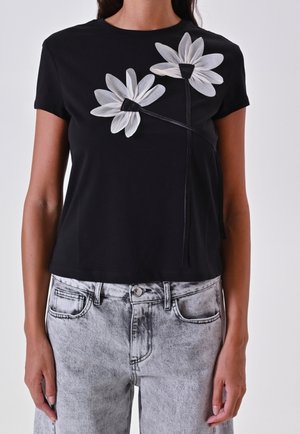 Femme portant un haut noir à manches courtes avec deux grandes fleurs de marguerite blanches et un jean gris délavé.