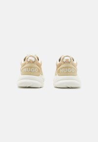 Beige i bijele sportske cipele s teksturiranim gornjim dijelom, s istaknutim logotipom na peti, okruglim vezicama i mekanim đonom.