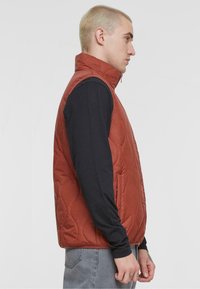 Quilted, rust-colored vest with high collar, side pockets, and zipper closure, layered over a black long-sleeve shirt.