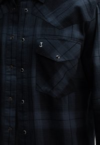 Zwart en donkerblauw geblokt overhemd met knoopsluiting, voorzien van zilveren drukknoopjes en een linkerknoopzakje met een geborduurd 'J'.