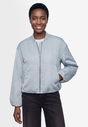 Uśmiechnięta kobieta w jasnoniebieskiej kurtce typu bomber z zamkiem błyskawicznym, czarnych spodniach, stojąca z jedną ręką w kieszeni na jednolitym tle.