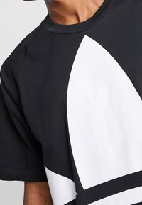 Person som bär en svart crewneck-t-shirt med en stor vit abstrakt geometrisk form på framsidan, närbild av axel- och bröstområde.