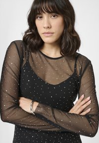 Donna con capelli scuri di lunghezza aderente che indossa un top trasparente nero con punti argentati e un braccialetto argento, le braccia incrociate di fronte a uno sfondo bianco.