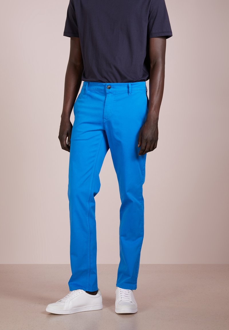 Homme portant un pantalon bleu vif, un t-shirt noir à manches courtes et des baskets blanches, debout devant un fond uni.