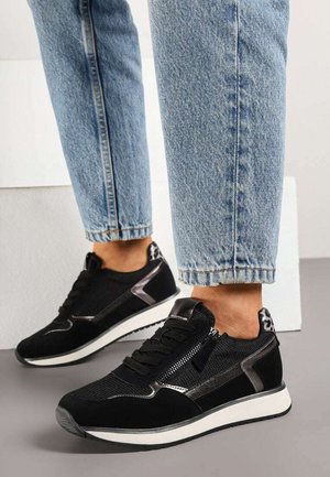 Baskets noires avec semelles blanches et détail de fermeture éclair portées avec un jean bleu clair retroussé sur un sol gris.