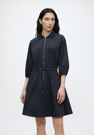 Žena s dlhými tmavými vlasmi, oblečená v čiernych šatách so zipsom, s tromi štvrťovými rukávmi a sťahovacím pásom v páse, stojaca pred jednoduchým pozadím.