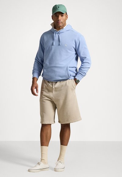 Sweat à capuche bleu clair avec une poche avant, short en coton beige, baskets blanches et casquette verte ; tenue de sport décontractée.