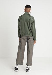 Olivgrön bomberjacka med vit kantning, elastisk nederkant och långa ärmar, kombinerad med grå rutiga byxor och svarta sneakers.