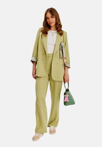 Světle zelený dvouřadý blejzr s odpovídajícími kalhotami. Bílý top pod ním. Drží zelenou kabelku s růžovým psím přívěskem. Béžové tenisky.