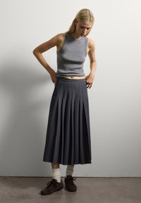 Giovane donna che sistema una gonna midi a pieghe blu navy, indossando un top senza maniche grigio, calze bianche e sneakers marrone scuro, in piedi di fronte a un muro bianco semplice.