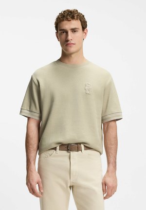 Ung mand stående iført en beige kortærmet skjorte med et lille broderet logo og lyse bukser med et brunt bælte.