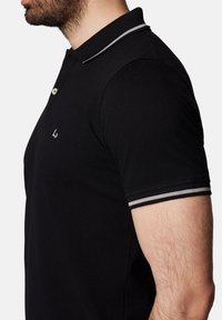 Czarna koszulka polo z teksturowanej tkaniny, z zapięciem na trzy guziki, z białymi i szarymi paskami na kołnierzyku i rękawach.