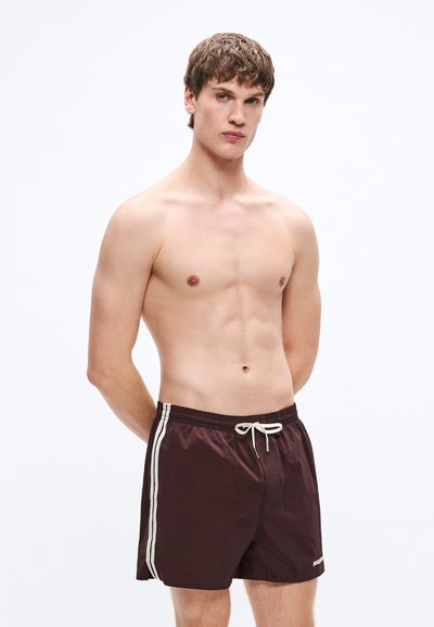 Pantalones cortos de baño marrones con rayas blancas laterales, cintura elástica, cordón ajustable y tejido liso y ligero. El modelo posa con las manos detrás de la espalda.