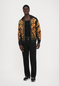 Homme portant un sweat à capuche zippé noir et or, un débardeur noir, un jean noir et des baskets noir et blanc, se tenant contre un fond blanc.