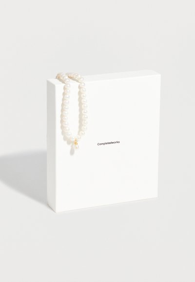 Σειρά από λευκές πέρλες με χρυσό κούμπωμα, τοποθετημένη πάνω σε ένα λευκό κουτί με την ετικέτα «Completedworks» σε ένα απλό φωτεινό φόντο.