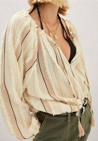Blouse légère à rayures en crème et beige, avec une finition texturée, un décolleté ouvert et un détail de cordon à frange. Associée à un pantalon vert olive.