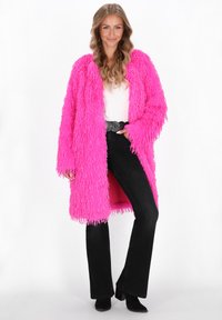Glunderende vrouw met lang golvend haar, gekleed in een felroze fluffy jas, een witte top, zwarte flared broek, een zwarte riem en zwarte hoge laarzen.