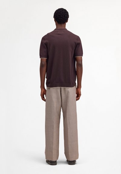 Mann mit kurzen, lockigen Haaren steht mit dem Rücken zur Kamera und trägt ein dunkelbraunes, kurzärmliges Strickshirt sowie hellbeige, weit geschnittene Hosen.