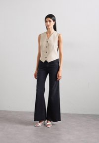 Beige linen vest with a fitted design and five black buttons, paired with flared black denim jeans and white open-toe heels.
