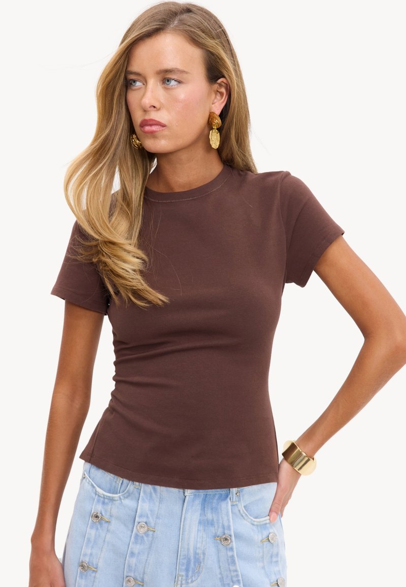 Femme aux longs cheveux blonds portant un t-shirt marron ajusté à manches courtes, une jupe en denim bleu clair à boutons, des boucles d'oreilles en or et un bracelet en or.