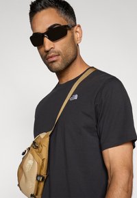 T-shirt noir, sac bandoulière beige avec logo, et lunettes de soleil foncées. Le sac est équipé de fermetures éclair et de bretelles ajustables. Design simple et fonctionnel.