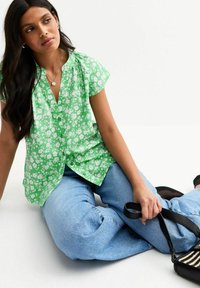 Blouse verte à manches courtes ornée de fleurs, avec des boutons à l'avant, associée à un jean taille haute bleu clair et un sac noir à rayures.