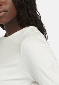 Camiseta blanca de manga larga hecha de material suave; cuenta con cuello redondo y un logo bordado en la manga izquierda. Textura suave.