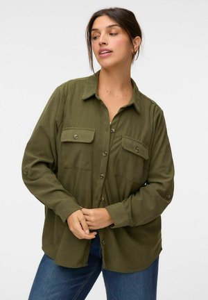 Femme portant une chemise boutonnée vert olive avec des poches poitrine et un jean bleu, debout devant un fond blanc uni.