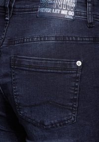 Dark denim jeans feature a textured surface with a back pocket, polished metal button, and a branded label near the waistline.