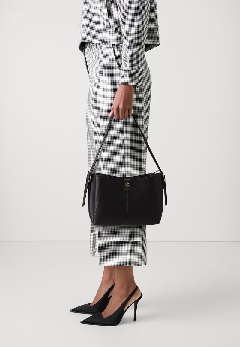 Personne en costume à carreaux noir et blanc tenant un sac à main en cuir noir, portant des escarpins slingback noirs à bout pointu, debout sur un sol blanc.