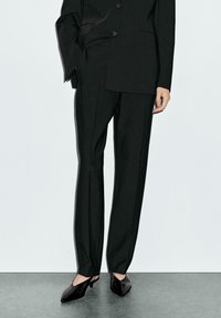 Black tailored suit with a structured silhouette, long trousers, slim fit, and smooth texture. Paired with black pointy-toed heels.