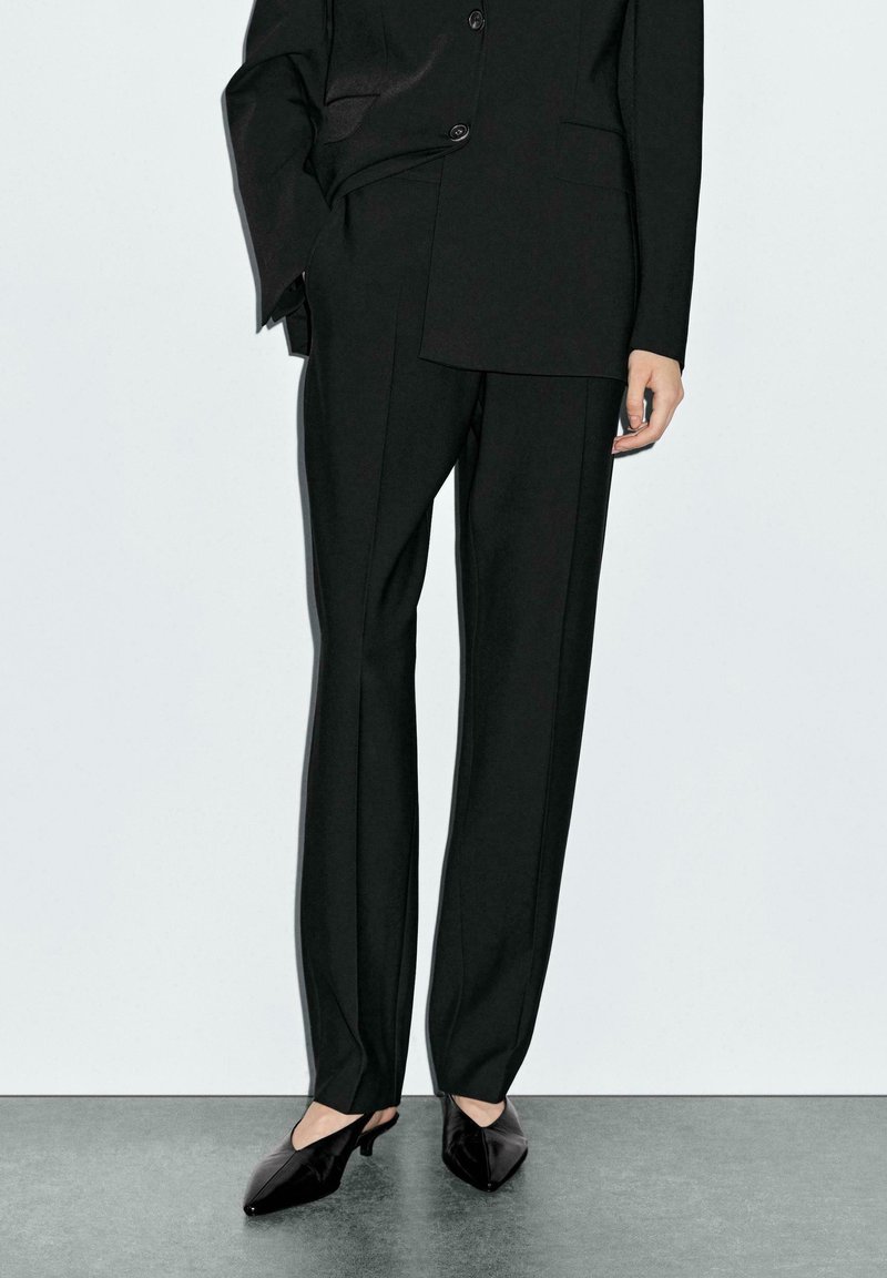 Zwart op maat gemaakt pak met een gestructureerde silhoutte, lange broek, slim fit en een soepele textuur. Gecombineerd met zwart puntig geschoeide hakken.