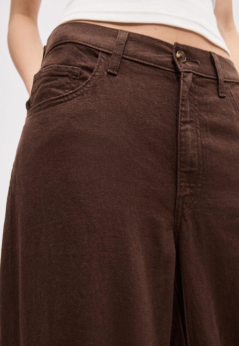 Close-up of brown high-waist pants with front pockets, button, and zipper, worn with a tucked-in white top.