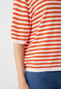 Pomarańczowy i biały sweter w paski z krótkimi rękawami, żebrowanym kołnierzykiem i dołem. W połączeniu z niebieskimi dżinsami, prezentującemu swobodny styl.
