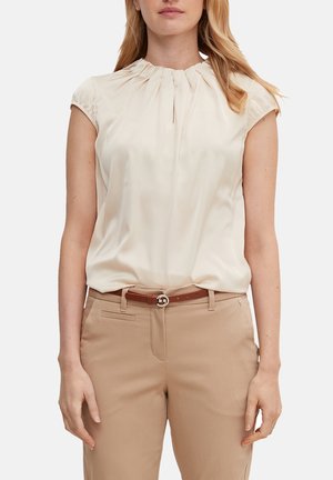 Femme portant un chemisier crème à manches courtes plissé, rentré dans un pantalon beige avec une ceinture marron, debout devant un fond uni.