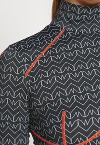 Svart rash guard med ett vitt zig-zag mönster, hög krage och kontrasterande orange sömmar längs kanterna. Slätt, elastiskt material.