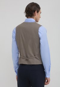 Braunes Stoffwesten mit strukturiertem Design, Rückengurtdetail, kombiniert mit einem hellblauen Langarmhemd. Texturiertes Material sichtbar.