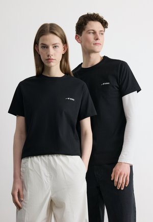 Νεαρή γυναίκα και άνδρας που φορούν μαύρα μπλουζάκια με το λογότυπο ARKK, στέκονται μπροστά σε απλό λευκό φόντο, η γυναίκα φοράει λευκό παντελόνι, ο άνδρας πουκάμισο με στρώσεις.