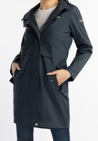 Žena v tmavě modré kapucí s dešťovou bundou, se zipem a patentky, má ruce v předních kapsách, má na sobě bílé tričko a modré džíny.