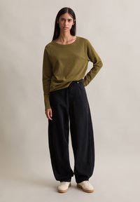 Olivenfarget genser med raglanermer, parret med løse svarte bukser. Modellen har på seg lysfargede joggesko. Enkel, minimalistisk design.