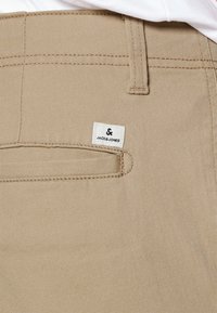 Beige bomullbyxor med en bakficka, som har en vit etikett märkt "JACK&JONES." Strukturerad tyg med synliga sömdetaljer.