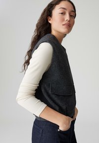 Gilet senza maniche in lana grigia con due tasche frontali, indossato sopra una camicia a maniche lunghe crema a costine, abbinato a jeans in denim scuro.