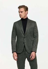Blazer gris oscuro con un sutil patrón de espiga, dos botones y solapas de muesca, combinado con pantalones ajustables a juego y un cuello alto negro.