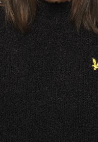 Maglione nero con un lavorazione a maglia testurizzata, caratterizzato da un piccolo logo di un'aquila gialla sul lato sinistro. La vista ravvicinata mette in risalto il tessuto e i dettagli.