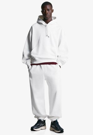 Jeune homme portant un hoodie blanc surdimensionné et un pantalon de survêtement assorti avec des baskets noires et blanches, se tenant avec les mains dans les poches.