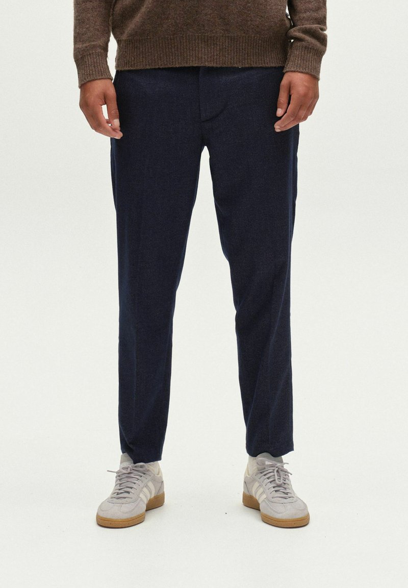 Sötétkék, egyenes szabású nadrág, sima textúrával. Világosszürke sportcipőkkel és barna pulóverrel viselve.