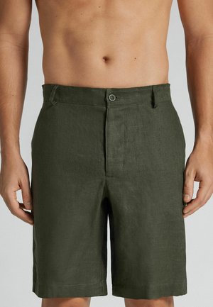 Mandligt torso iført olivengrønne knapshorts med bæltestropper, stående med arme afslappet mod en ensfarvet lys baggrund.