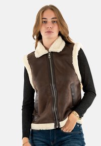 Gilet en cuir marron avec col en shearling crème, fermeture éclair, poches latérales et texture contrastante. Porté par-dessus un haut à manches longues noir.