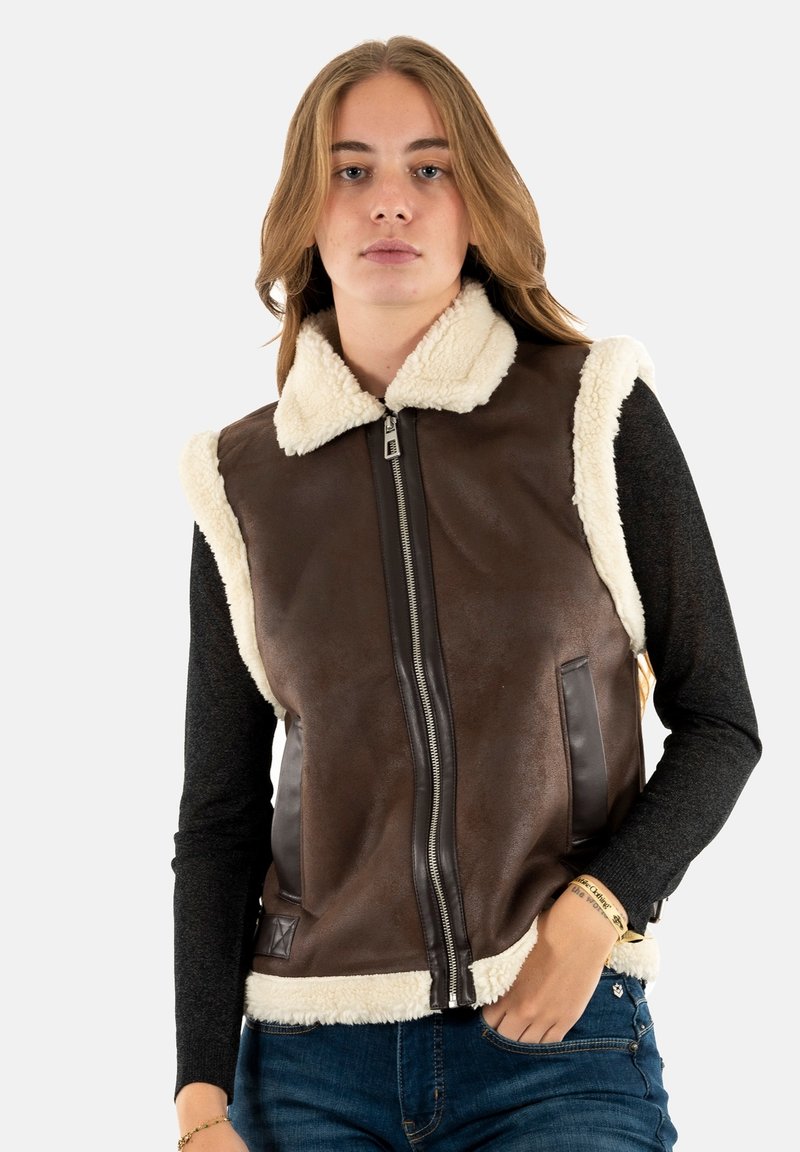 Gilet en cuir marron avec col en shearling crème, fermeture éclair, poches latérales et texture contrastante. Porté par-dessus un haut à manches longues noir.